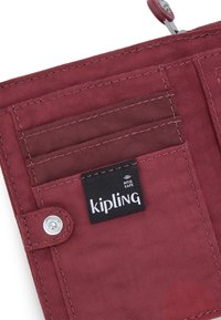 Czerwony portfel Kipling wykonany z trwałego materiału, z wieloma kieszeniami na karty, zapięciem na zatrzask oraz etykietą ochrony RFID. Teksturowana powierzchnia.