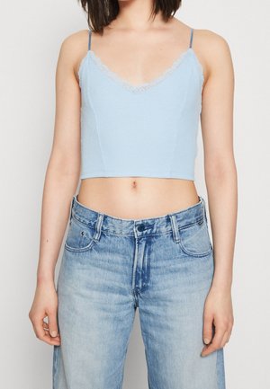 Kobieta ubrana w jasnoniebieski krótki top na ramiączkach z koronkowym wykończeniem oraz wysokiej talii wyblakłe niebieskie dżinsy, stojąca na jednolitym tle.