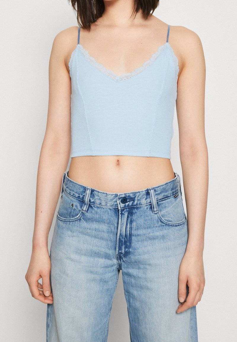 Femme portant un caraco court bleu clair avec des bordures en dentelle et un jean bleu délavé taille haute, debout devant un fond uni.