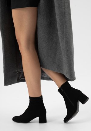 Benen met zwarte enkellaarzen met hak en zijritsen, met een grijze jas over een zwarte korte broek gedrapeerd.