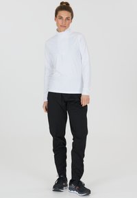 Weißes langärmeliges Baselayer mit halbem Reißverschluss-Kragen, kombiniert mit schwarzen tapered Pants und schwarzen Sportschuhen mit Akzenten. Glatte Textur.