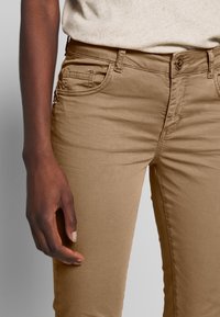 Närbild av en person i beiga byxor och en ljusbeige topp, med vänster arm avslappnat vid sidan mot en neutral bakgrund.