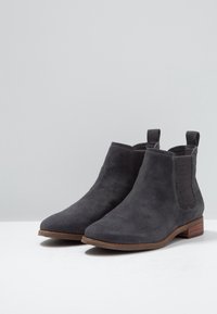 Par de botas Chelsea de ante gris oscuro con paneles elásticos en los laterales y tacones bajos de madera, exhibidas sobre una superficie blanca con un fondo gris claro.