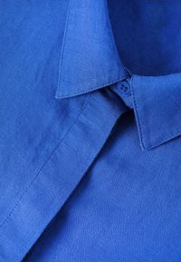 Gros plan sur le col d'une chemise en lin bleu avec un bouton fermé, montrant la texture du tissu et les coutures détaillées.