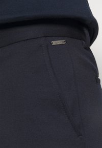 Tamno plave hlače s glatkom teksturom, koje imaju prednju džep i metalnu oznaku "Jack & Jones" na pojasu.