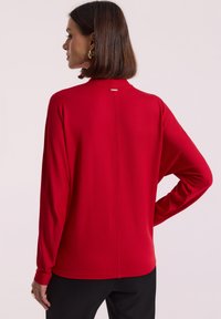 Kobieta z brązowymi włosami do ramion, nosząca złote kolczyki w kształcie kół oraz czerwoną bluzkę z długimi rękawami, ukazana z tyłu z jedną ręką na biodrze.