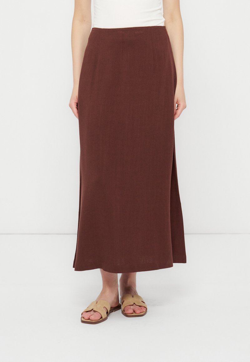 Femme portant une longue jupe droite marron avec des fentes latérales, associée à des sandales plates ouvertes beige, debout devant un fond uni.