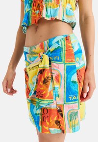 Falda envolvente de estampado colorido que presenta gráficos tropicales en azul, naranja y amarillo, hecha de un tejido ligero, atada a la cintura.