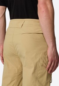 Beige cargo-housut sileällä pinnalla, joissa on takatasku ja sivuvetoketju, suunniteltu käytännöllisyyttä ja mukavuutta varten.