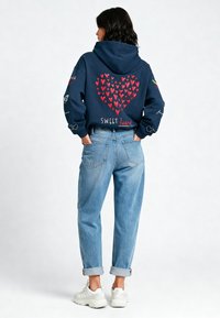 Donkerblauwe hoodie met een rood hartontwerp en de tekst "SWEET" op de achterkant. Gecombineerd met licht gewassen omgerolde jeans en witte sneakers.