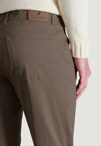 Persona con pantaloni marroni dotati di tasca posteriore e una patch in pelle con l'etichetta "Jeckerson", e una manica di un maglione a maglia color crema visibile.
