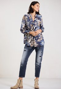 Langärmlige Bluse mit blauem Blumen- und Tierdruckmuster, kombiniert mit ausgebleichten blauen Jeans und beigen Knöchelstiefeln.