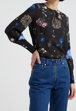 Frau trägt eine schwarze, langärmelige Bluse mit Blumenmuster und gerafften Manschetten sowie hochtaillierte blaue Jeans mit Reißverschluss vorne.