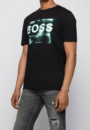 Zwart katoenen T-shirt met een witte en groene "BOSS"-grafiek op de voorkant, korte mouwen en een losse pasvorm. Gecombineerd met versleten grijze jeans.