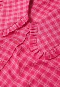 Chemise à carreaux rose avec un col volanté et une patte de boutonnage. Les boutons sont également roses. Tissu léger avec une texture douce, présentant un motif en quadrillage.