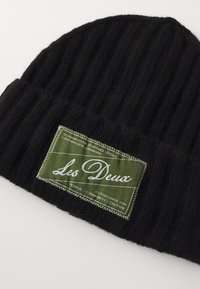 Bonnet tricoté noir côtelé avec une étiquette rectangulaire verte arborant un texte blanc et des détails de couture. Texture douce, forme ajustée.