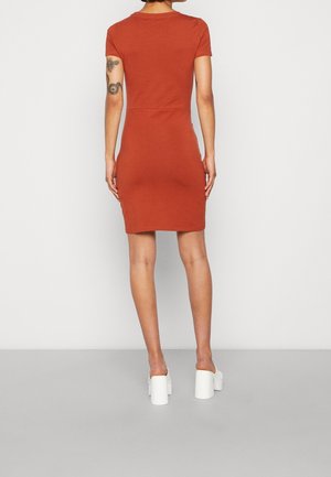Femme aux cheveux courts portant une robe cintrée à manches courtes couleur rouille et des talons compensés blancs, montrant un tatouage sur son bras supérieur droit.