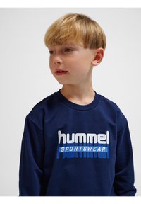 Felpa navy in cotone con collo rotondo. Presenta il logo "hummel" e "SPORTSWEAR" in bianco e blu sulla parte anteriore.