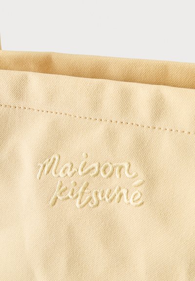 Tissu couleur crème avec texte brodé "Maison Kitsuné" en fil assorti sur une surface textile.