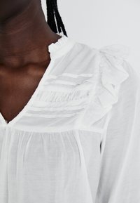 White blouse with ruffled accents along the neckline, featuring pleated details and a lightweight fabric. V-shaped collar design.