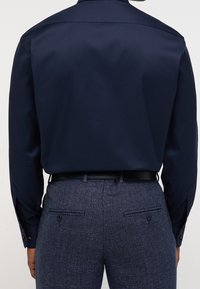 Tmavě modrá košile s dlouhým rukávem a hladkou texturou, vybavená klasickým límečkem a manžetami na knoflíky, v kombinaci s texturovanými tmavými kalhotami.