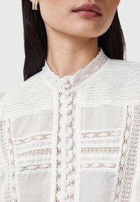 Camicia bianca con strisce testurizzate e dettagli in pizzo. Presenta un colletto alto ricamato e bottoni frontali. Tessuto semi-trasparente.
