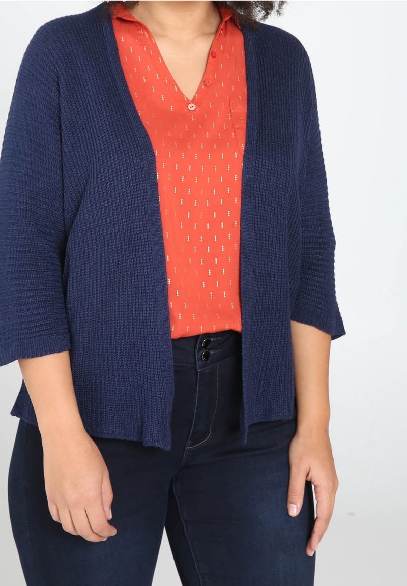 Cárdigan de punto azul marino con un corte relajado y mangas de tres cuartos, superpuesto a una camisa roja con cuello y pequeños patrones en blanco.