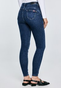 Žena u tamnoplavim uskim trapericama s džepovima na stražnjoj strani i crnim sandalama na petu, stoji ispred jednostavne svijetle pozadine.