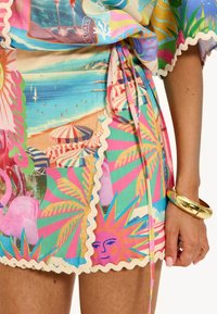 Étole de plage multicolore avec imprimés tropicaux, bords festonnés et taille nouée. Le matériau semble léger et aéré. Bracelet en or porté.