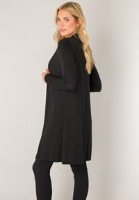 Zwarte jurk met lange mouwen en een losse pasvorm, gemaakt van zacht stof, met een rechte snit en iets uitlopende zoom. Draagt zwarte legging erbij.