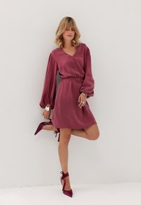 Różowa sukienka z długimi, puszystymi rękawami, dekoltem w kształcie litery V i materiałowym paskiem. Połączona z burgańskimi szpilkami z wiązaniem na kostce. Gładka faktura.