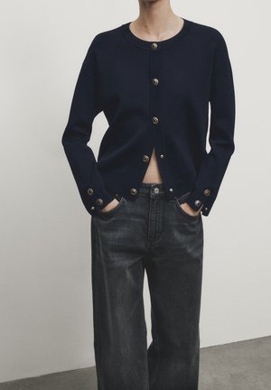 Personne portant un cardigan bleu marine boutonné avec des boutons métalliques et un jean noir taille haute, mains dans les poches, debout devant un fond neutre.