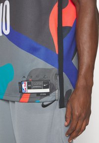Šedý sportovní dres s mnohobarevným abstraktním vzorem, s černým bočním švem, štítkem s logem Nike a autentickou NBA etiketou.
