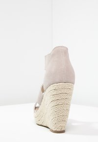 Sandal con zeppa beige con tacco platform in juta intrecciata e cinturino alla caviglia in suede, su superficie e sfondo bianchi.