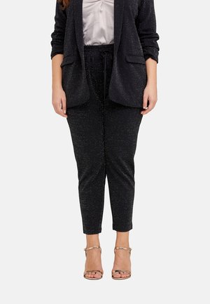 Femme portant un pantalon noir scintillant fuselé, un blazer assorti, un chemisier blanc froncé, et des talons ouverts métalliques à bride cheville, debout devant un fond blanc.