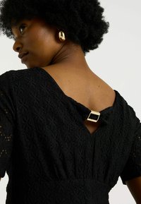 Femme aux cheveux courts et bouclés portant une robe noire texturée avec une boucle en or au dos du col et des boucles d'oreilles en or.