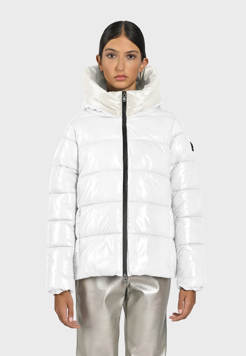 Dikke, glanzende witte jas met capuchon, voorzien van een zwarte rits en geribbelde manchetten. Draagt over metallic zilveren broeken met een gladde textuur.