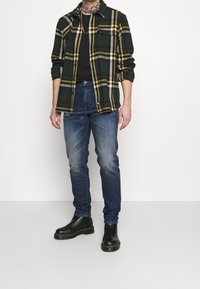 Camisa de franela a cuadros verde y amarilla sobre una camiseta negra, combinada con jeans azules descoloridos y botas negras de tobillo. Tejido texturizado, ajuste casual.