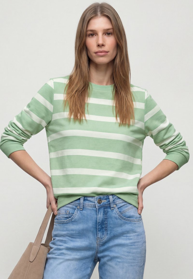 Femme aux cheveux longs châtain clair portant un pull rayé vert et blanc et un jean bleu, tenant un sac fourre-tout beige à son côté.