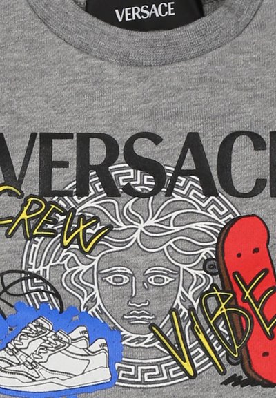 Felpa grigia con collo rotondo che presenta la scritta nera "VERSACE", una grafica centrale di un viso e accenti colorati tra cui sneakers e uno skateboard.