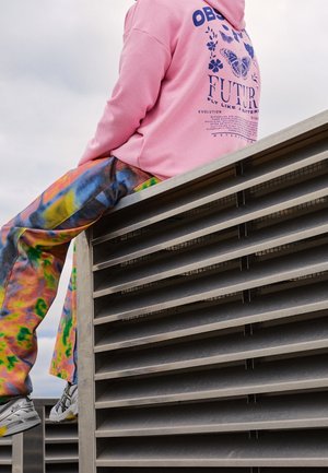 Persona con indosso una felpa con cappuccio rosa con testo farfalla e pantaloni tie-dye colorati seduta su una struttura metallica di ventilazione all'aperto.