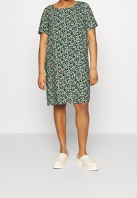 Kortärmad klänning med svart bakgrund och blommönster i gröna, vita och orange toner; avslappnad passform, knä-längd. Ljus beige träskor bärs.