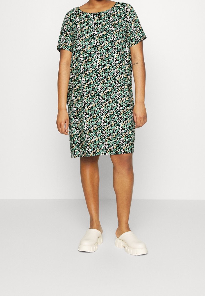 Kortärmad klänning med svart bakgrund och blommönster i gröna, vita och orange toner; avslappnad passform, knä-längd. Ljus beige träskor bärs.