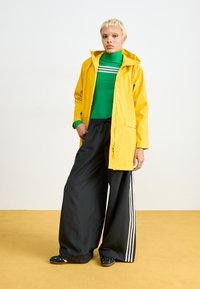 Capa de chuva amarela com capuz, blusa verde de mangas longas, calças largas pretas com três riscas brancas e sapatos slip-on com padrão azul.