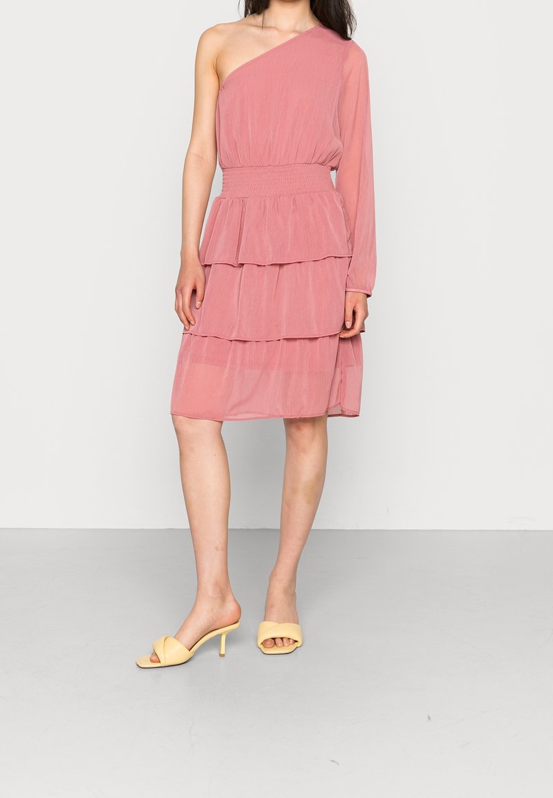 Femme dans une robe rose asymétrique à une épaule, à manches longues et à volants, portant des sandales jaunes à talons, debout devant un fond clair et uni.