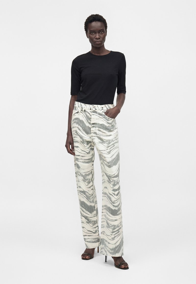 Frau steht und trägt ein schwarzes, figurbetontes Oberteil, grau-weiß gestreifte, hoch taillierte weite Hosen und schwarze offene High Heels.