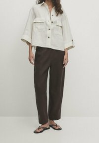 Beige linen cropped shirt with two chest pockets and brown buttons, paired with dark brown loose-fitting trousers and black flip-flops.