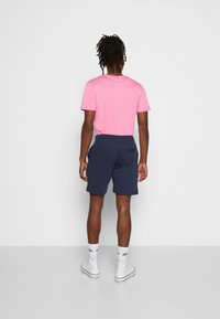 Rosa Kurzarmhemd, marineblaue Shorts mit Seitentaschen und weiße High-Top-Sneaker. Der Stoff wirkt weich und lässig im Schnitt.