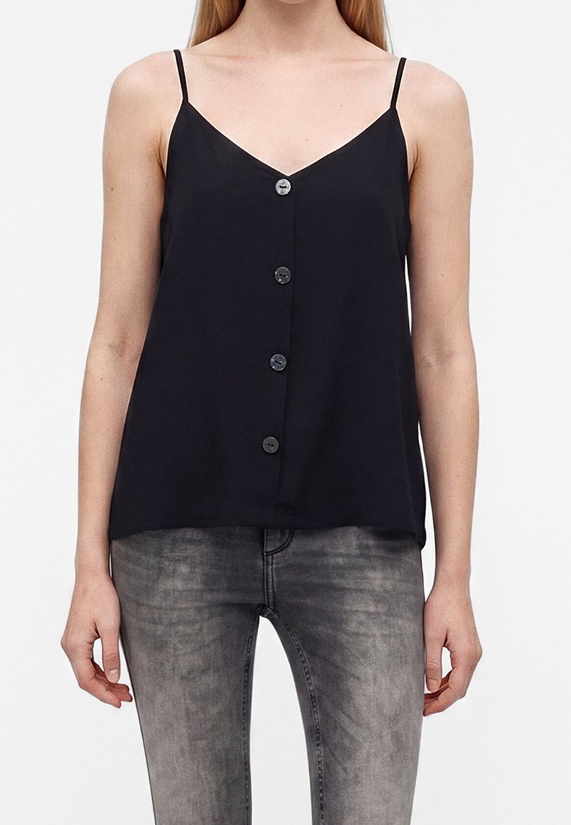 Femme portant un haut noir sans manches à boutons avec fines bretelles et un jean gris délavé, debout devant un fond uni.