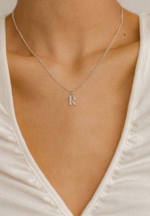 Silver necklace with a shiny, textured chain and a diamond-accented letter "R" pendant, worn against a textured white top.